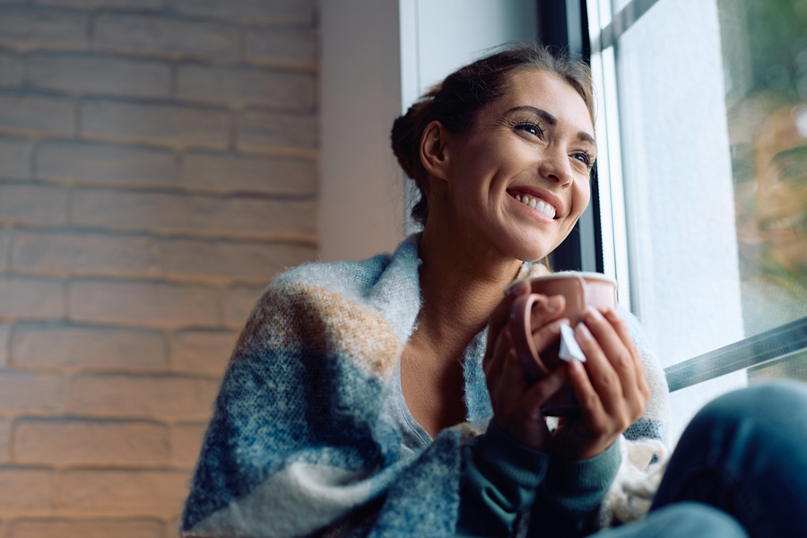 A woman smiling as she sits by a window with a hot drink