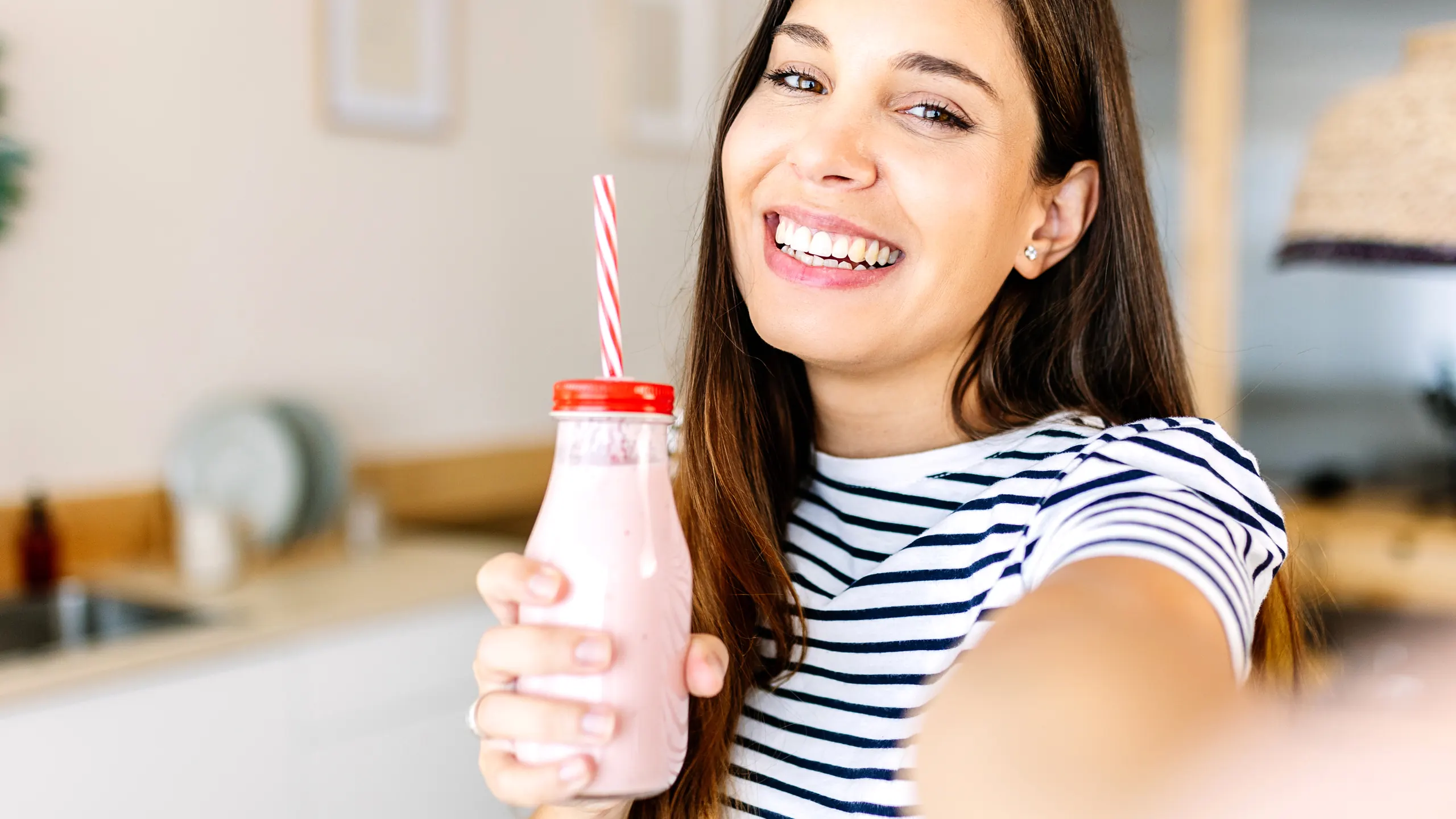 Smiling woman holding up a pink meal replacement shake in a glass bottle with a striped straw